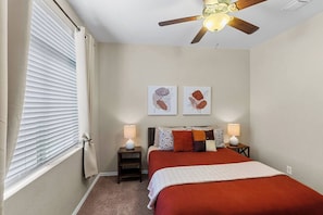 A bedroom with a bed, two bedside tables with lamps, a ceiling fan, and artwork on the wall.