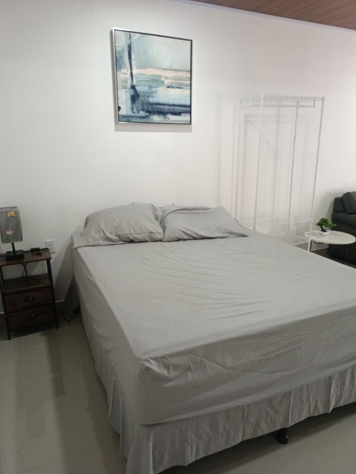 Un dormitori amb un llit, una taula de nit, una taula petita amb una planta, un quadre a la paret i una finestra amb cortines.