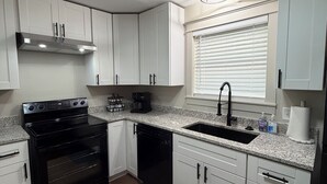 A compact kitchen with white cabinets, a black stove, a dishwasher, and a window with blinds. - Kimberly House (Mena)
