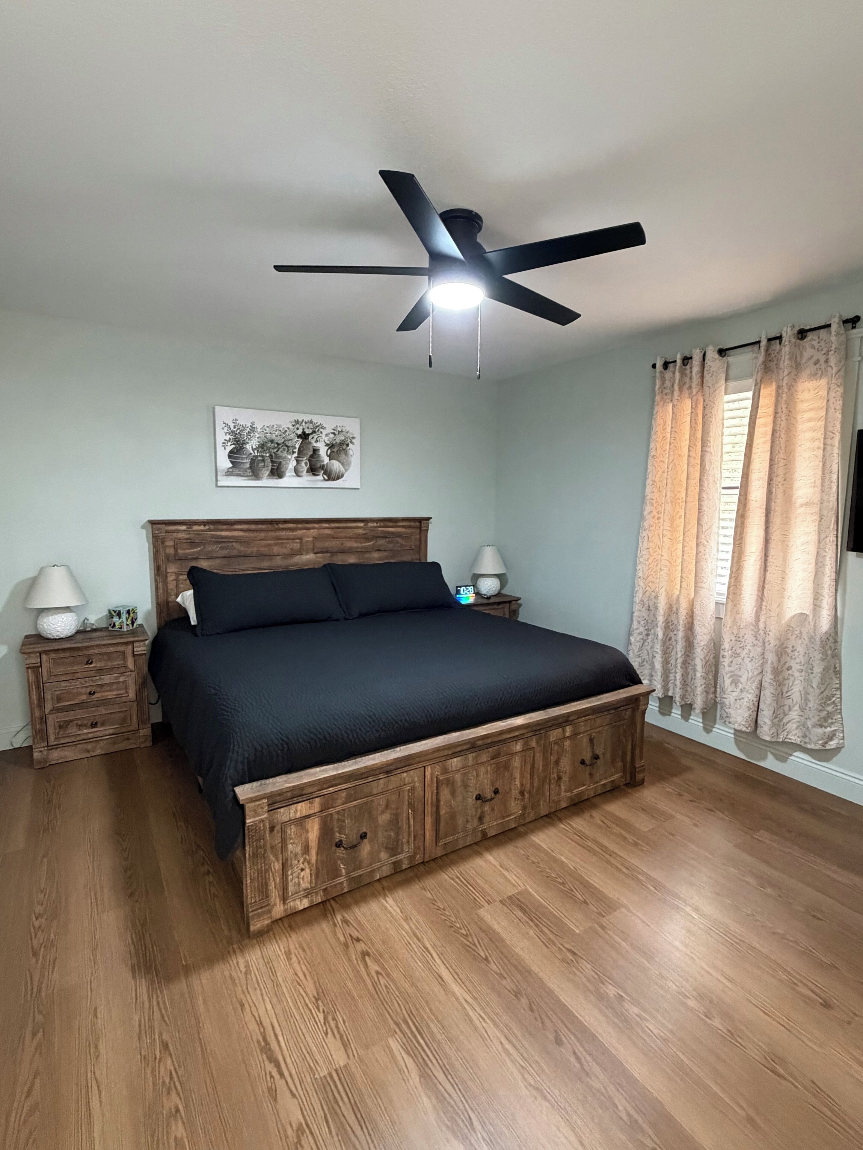 A bedroom with a wooden bed, a ceiling fan, a bedside table with a lamp, and a window with curtains.