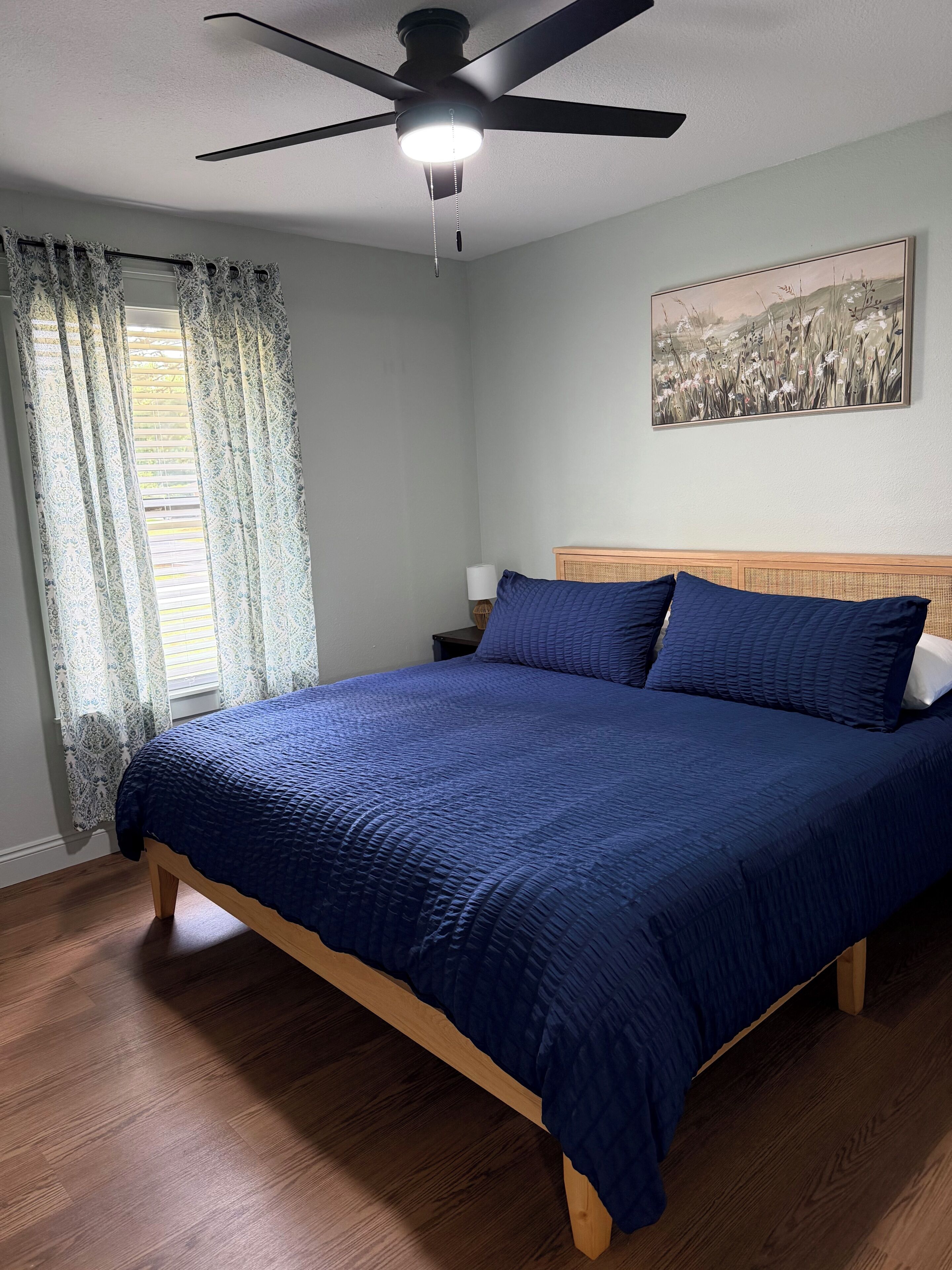 Una camera da letto con un letto, una ventola a soffitto, un comodino, una lampada e un quadro appeso al muro.