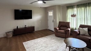 A living room with a ceiling fan, a flat-screen TV, a brown leather armchair, a brown sofa, a patterned rug, and a wooden coffee table. - Kimberly House (Mena)
