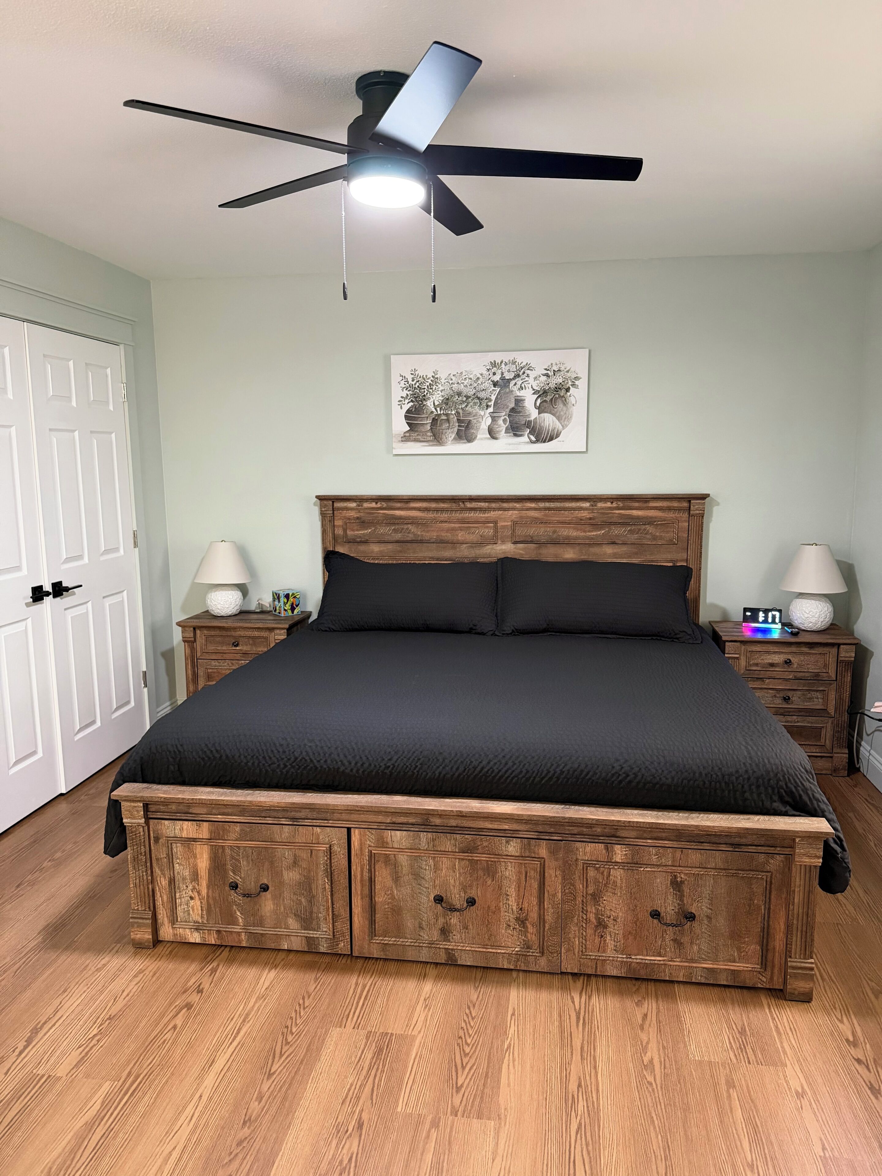 A bedroom with a bed, wooden headboard, bedside tables, a ceiling fan, and a wall-mounted artwork.