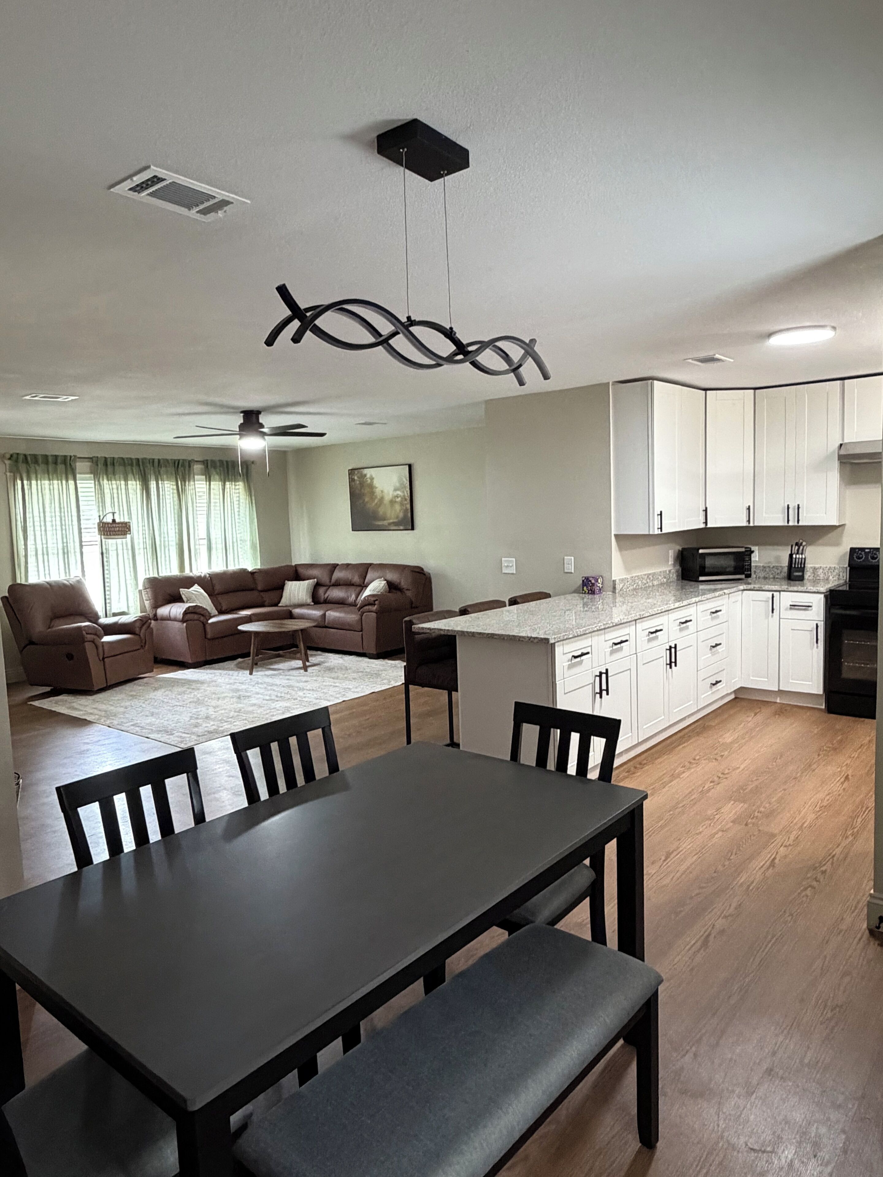 A modern living room with a brown leather sofa, a dining table with chairs, a kitchen with white cabinets, and a ceiling fan.