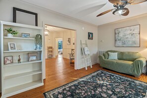 Ein Zimmer mit Holzboden, einem grünen Sofa, einer weißen Regaleinheit mit Dekoration und einem Deckenventilator, sowie einem großen abstrakten Gemälde.
