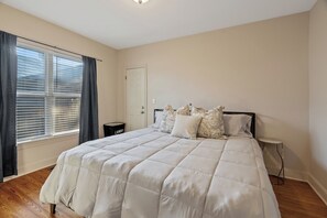 Ein Schlafzimmer mit einem Bett, einem Nachttisch, einem Fenster mit Vorhängen, einer Tür und einem kleinen Tisch.