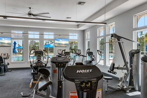 Ein Fitnessstudio mit verschiedenen Trainingsgeräten, großen Fenstern und einem Deckenventilator.