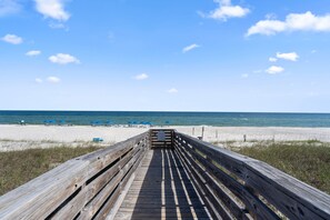 Ein hölzerner Steg führt zu einem Strand mit blauen Sonnenschirmen und Liegestühlen.