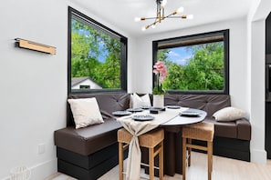 A modern dining area with a round table, two brown sofas, and large windows with a view of greenery.
