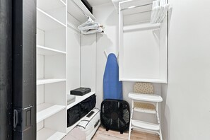 A compact room with a white wardrobe, a small ironing board, a fan, and a storage bench.