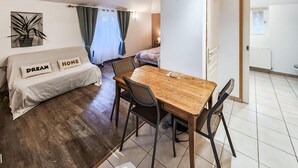 Ein Zimmer mit einem Bett, einem Holztisch mit Stühlen, einer Zimmerpflanze und einem Fenster mit Vorhängen.
