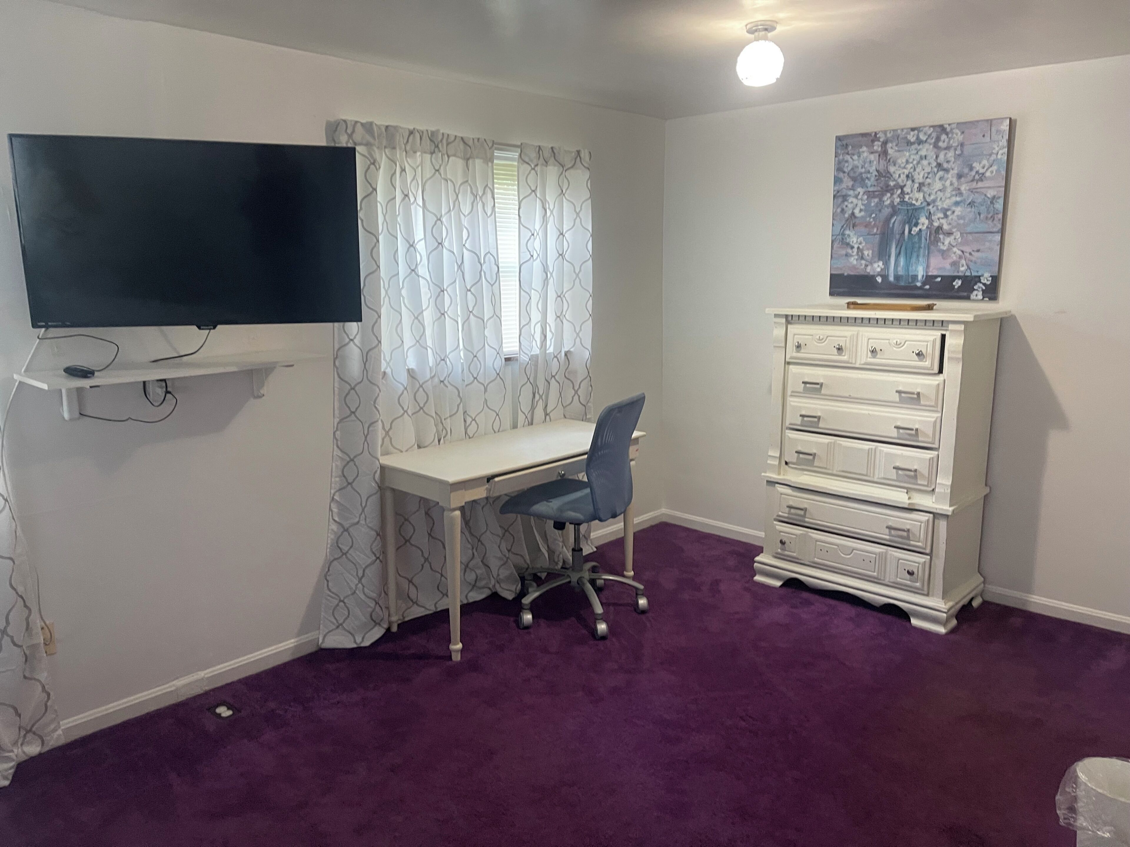 A room with a desk, chair, TV, dresser, and a painting on the wall.
