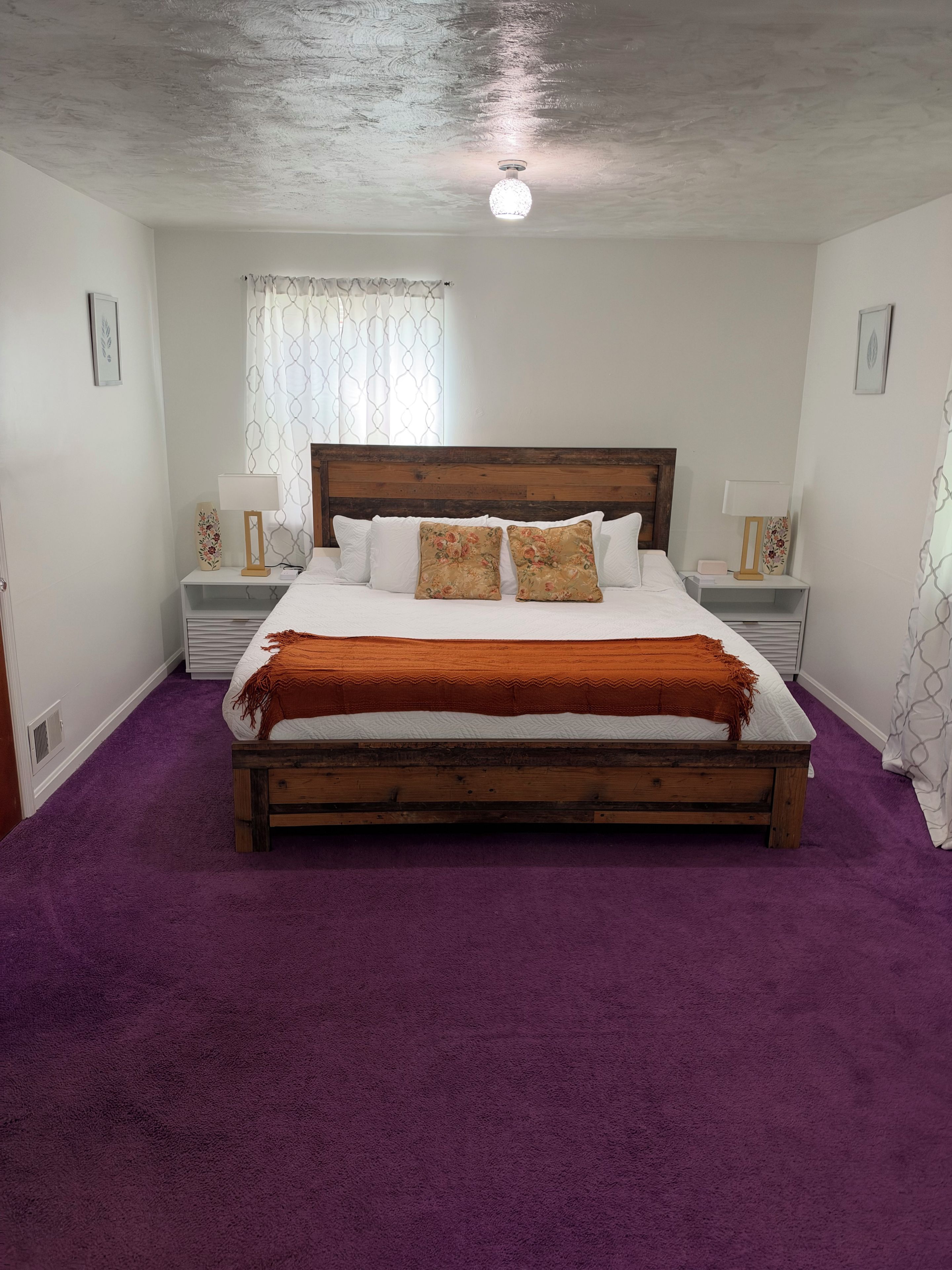 Un dormitorio con cama de madera, alfombra morada, paredes blancas y una lámpara en el techo.
