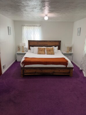 Une chambre à coucher avec un lit en bois, une moquette violette, des murs blancs et un luminaire au plafond.