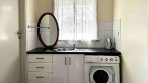 A compact kitchen with a sink, a washing machine, a mirror, and a window with lace curtains. - Ringwood 4BR House near Eastland + Parking (Ringwood)