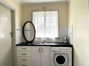 A compact kitchen with a sink, a washing machine, a mirror, and a window with lace curtains.