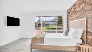 A bedroom with a bed, a TV mounted on the wall, a window with a view of the outside, and a brick wall. - Callow Place 10 - town location, relaxation, easy coastal living, Wifi (Bermagui)