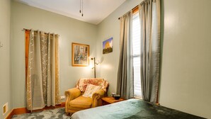 Una stanza con un letto, una sedia, un tavolo, una lampada e due finestre con tende.