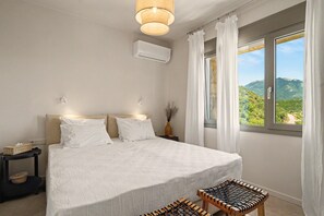 A bedroom with a bed, bedside tables, a nightstand, a window with a view, and an air conditioning unit. - Isichia Apartment with Sea and Taygetos Mountain Views (West Mani)