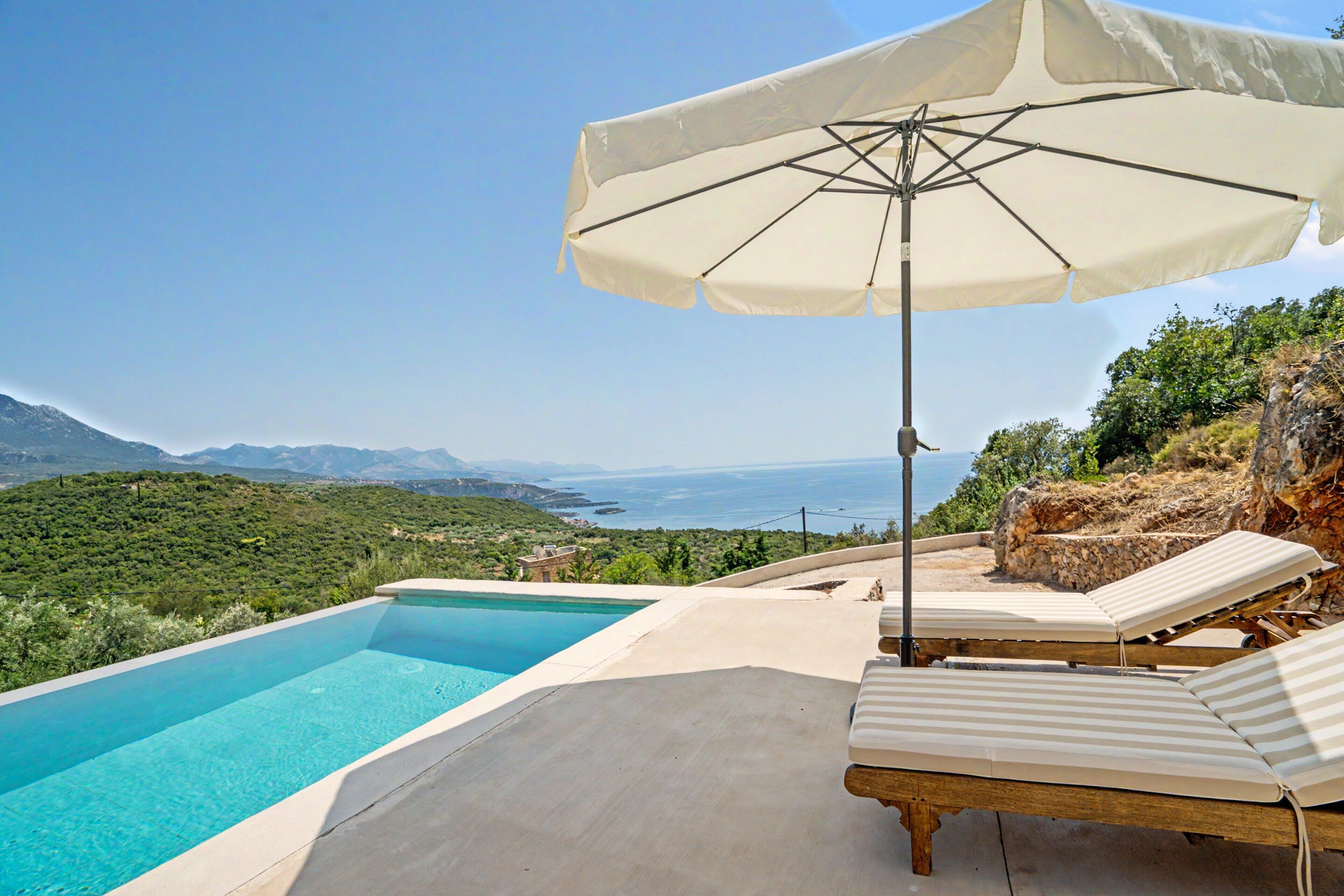 A pool with a view of the sea and mountains, a lounge chair, and an umbrella.