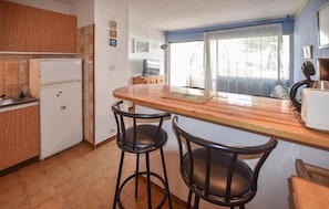 A compact kitchen with a refrigerator, a wooden countertop, two bar stools, and a window with curtains. - 1 bedroom beautiful apartment in Le Grau-du-Roi (Le Grau-du-Roi)