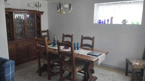 Ein Esszimmer mit Holztisch, vier Stühlen, einer Chandelüster, einem Fenster mit Dekoration und einem Schrank.