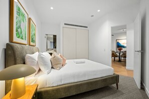 Quarto moderno com cama grande, mesa de cabeceira com luminária, guarda-roupa e área de jantar visível através de uma porta aberta.