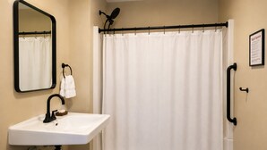 A bathroom with a white sink, a black faucet, a shower curtain, a towel rack, a handheld showerhead, and a framed informational sign. - Comfy & Pet Friendly Stay Near Cooperstown - ADA (East Springfield)