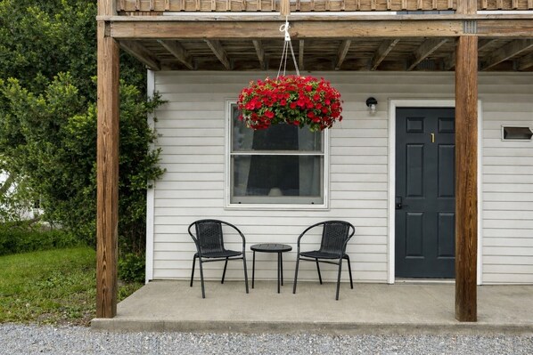 Une maison avec un porche, deux chaises, une jardinière suspendue et une porte.