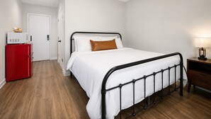 Un dormitorio con cama de estructura metálica, ropa de cama blanca, un electrodoméstico rojo y una mesita de noche con lámpara.