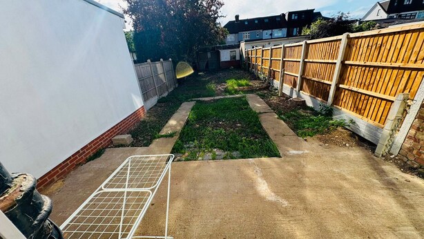 Ein Garten mit einem Betonweg, einem Metallgrill und einem Holzzaun.