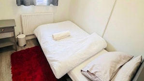 Ein kleines, ordentlich bezogenes Bett mit weißer Bettdecke und Kissen, einem roten Teppich, einem weißen Heizkörper und einem grauen Nachttisch mit Schublade.