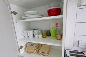 Um armário de cozinha com prateleiras brancas contendo diversos utensílios, incluindo pratos, copos e uma jarra de vidro.