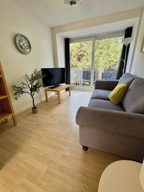 Ein Wohnzimmer mit grauem Sofa, einem Fernseher auf einem Holzboden, einer Topfpflanze und einem Balkon mit Blick auf Bäume.