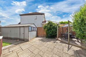 Ein Garten mit Terrasse, Gartenhaus, Backsteinmauer und hohem Gebäude im Hintergrund.