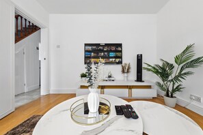 Ein modernes Wohnzimmer mit weißer Wand, einem Wand-Fernseher, einem runden Tisch mit Vase und einer Topfpflanze.