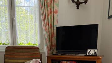 Sala de estar com televisão, estante de jogos de tabuleiro, cortina florida e janela com vegetação lá fora.