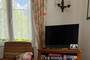 Una sala de estar con televisor, una estantería llena de juegos de mesa, una cortina con motivos florales y una ventana que da a zonas verdes.