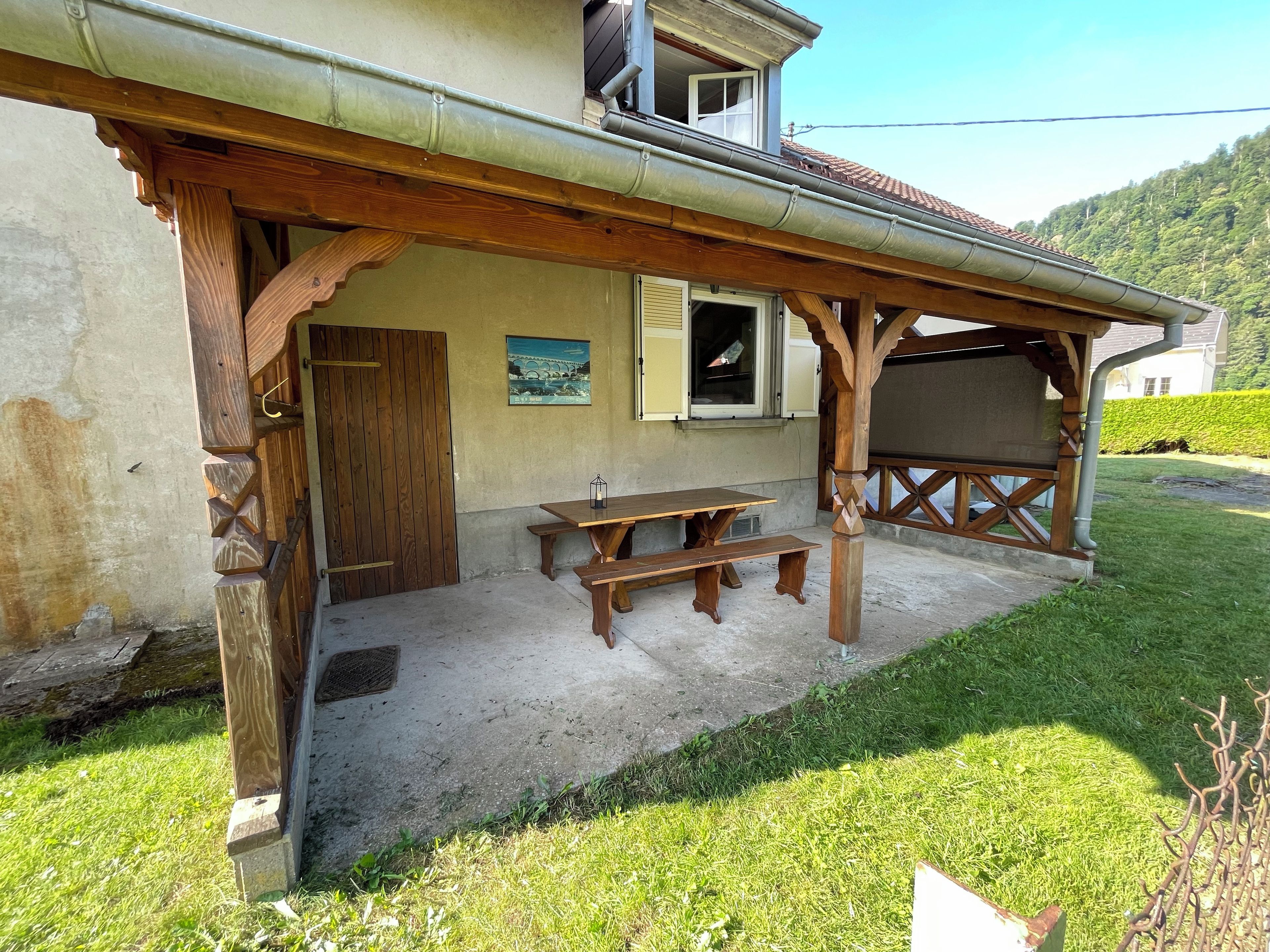 Eine Holz-Pergola mit Picknicktisch, Bänken, einer Tür und einem Fenster mit Fensterläden.