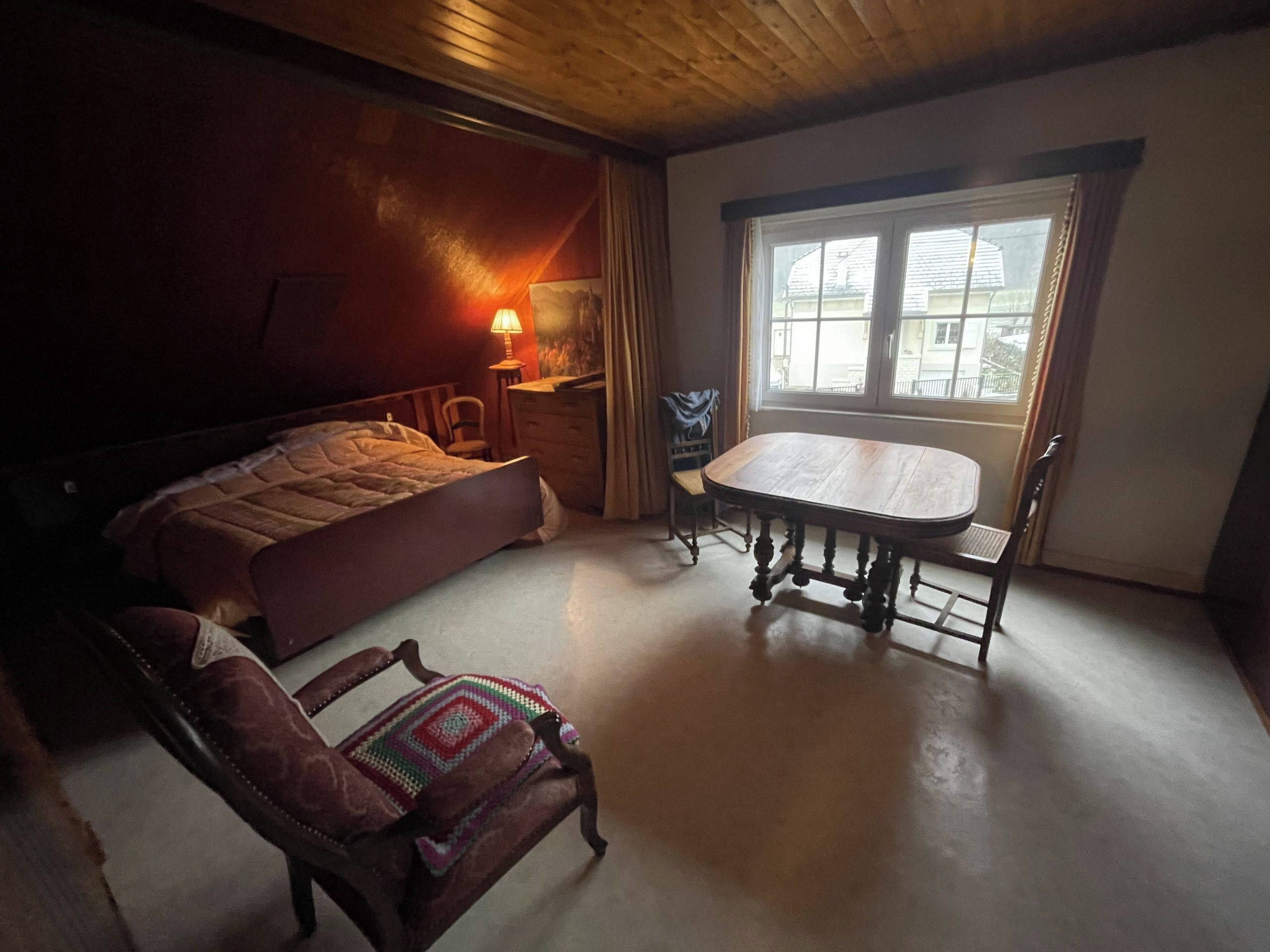 Une chambre à coucher avec un lit, une table en bois, une chaise, une lampe et une fenêtre.