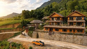 Un edificio de madera de varios pisos con balcones, ubicado en una ladera con vista a un río y montañas.