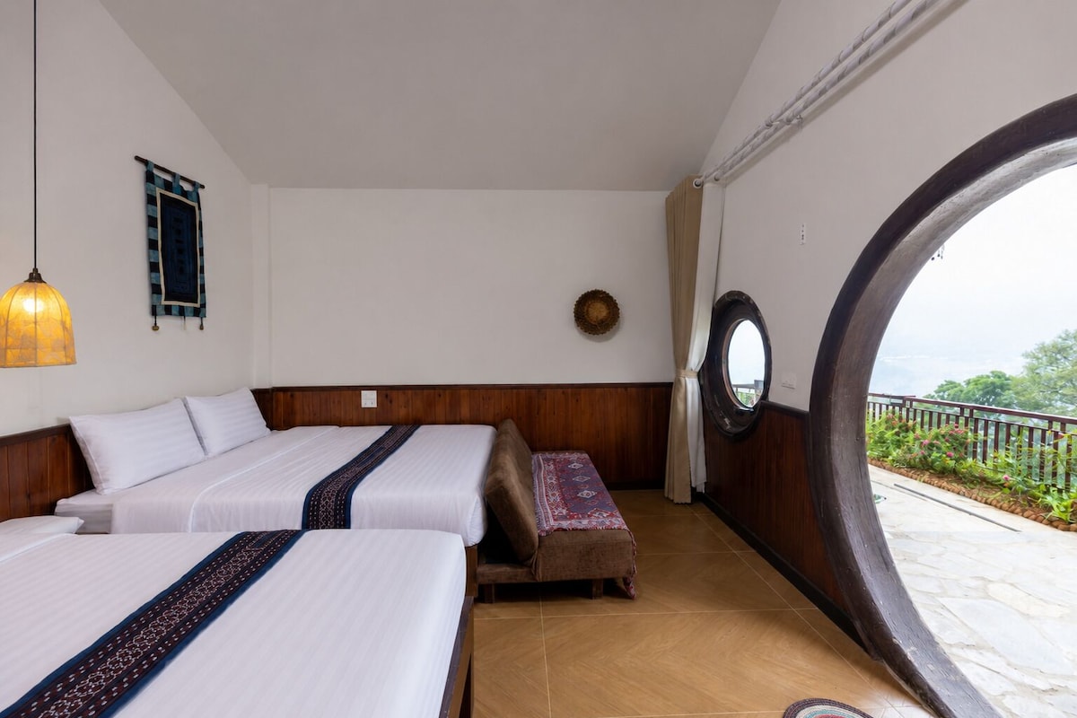 A room with two beds, a wooden headboard, a hanging light fixture, a round mirror, and a view through an arched window.