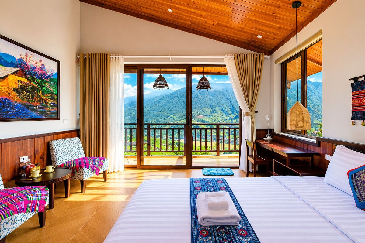 A hotel room with a bed, a desk, a chair, a table with a lamp, a painting, and a balcony with a mountain view.