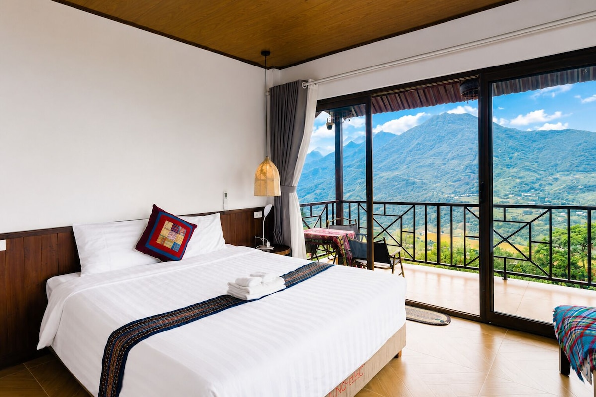 A hotel room with a large bed, a view of mountains, and a balcony with a table and chairs.
