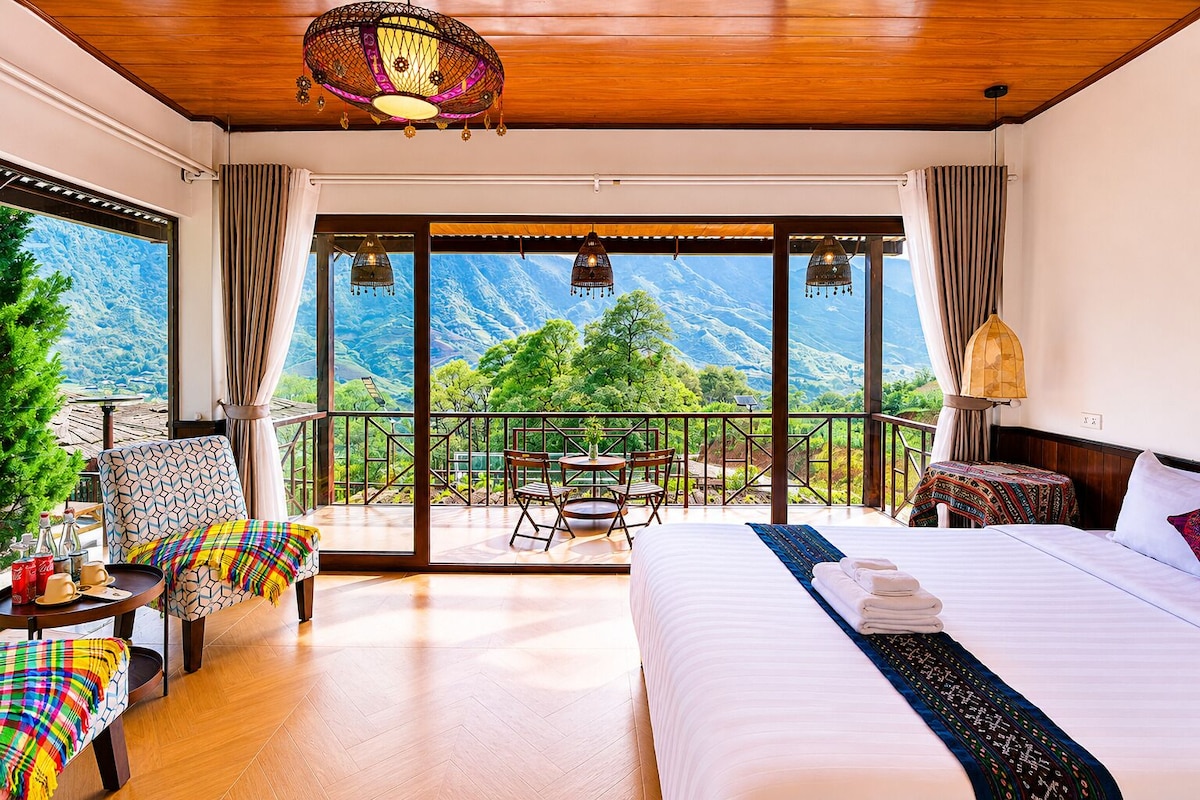 A hotel room with a bed, a chair, a table, and a view of mountains.