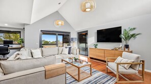 A modern living room with a sectional sofa, a coffee table, a TV, and a large window with a view. - Pomora Family Escape Close to Beach & Golf (Torquay)