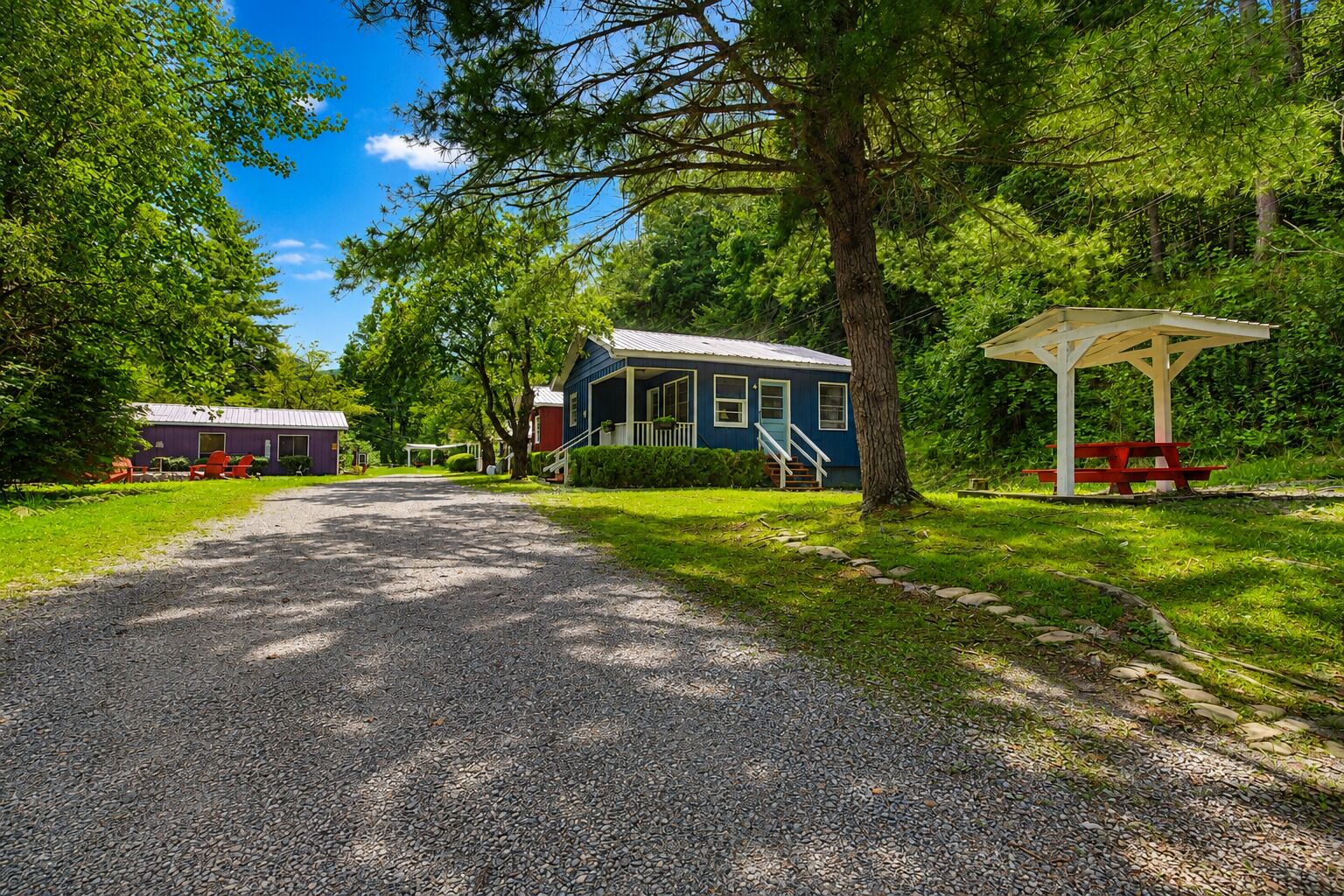 A gravel driveway leading to a blue house with a porch, surrounded by trees and a picnic table.
