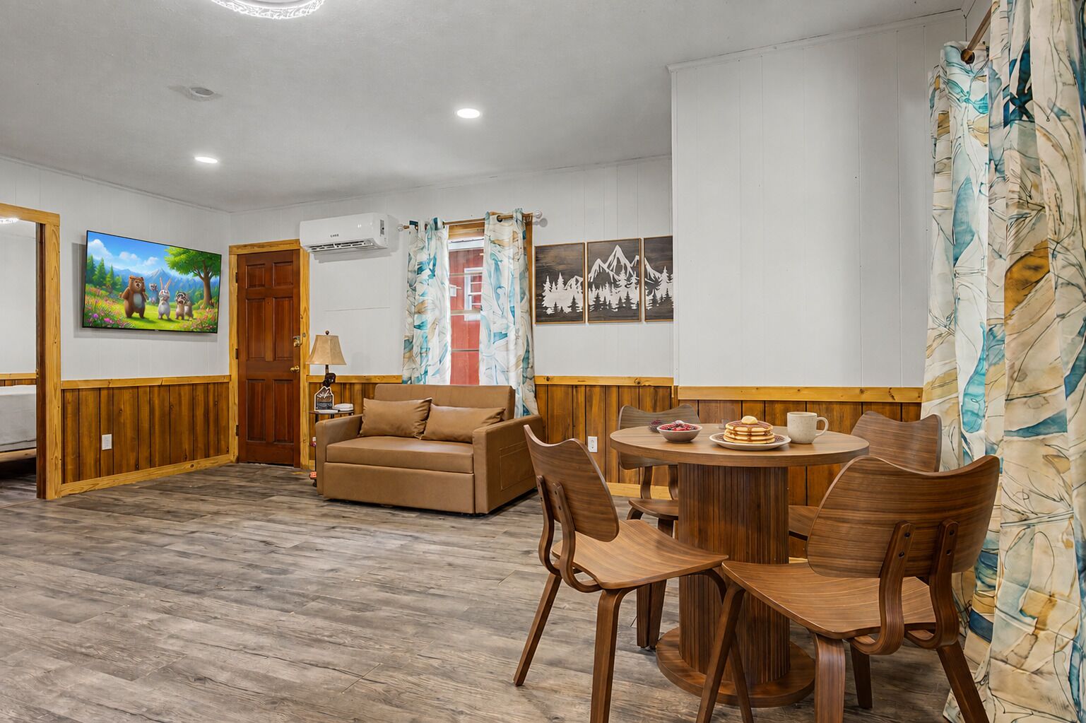 A room with a wooden floor, a sofa, a dining table with chairs, a TV, and a wall with framed pictures.