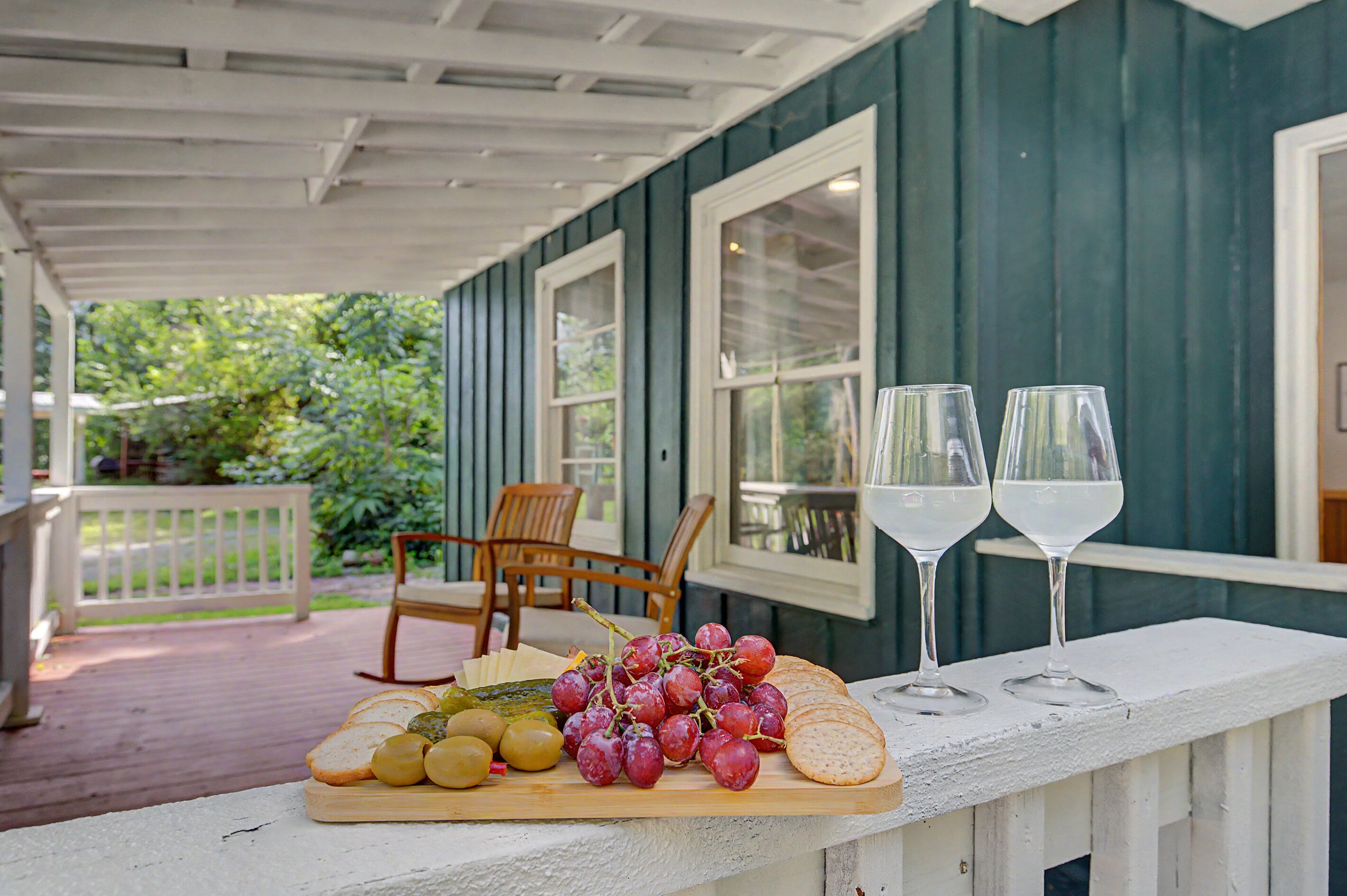 A wooden table with a cheese board, grapes, and crackers on a porch. Two wine glasses are placed on the table.