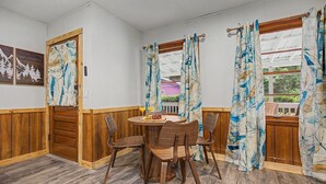 Un área de comedor con una mesa y sillas de madera, una pequeña mesa redonda con frutas y bebidas, y una vista a través del ventanal.